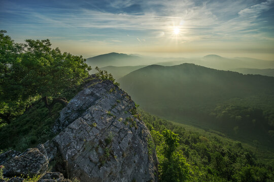 View of jagged grey rocks meeting lush green foliage, overlooking a misty mountain range under a bright sun, on Vysoka mountain, Kuchyna, Bratislava Region, Slovakia.
