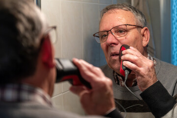 An elderly man wearing glasses shaves his face with an electric razor while looking in the bathroom mirror, showing daily grooming at home.