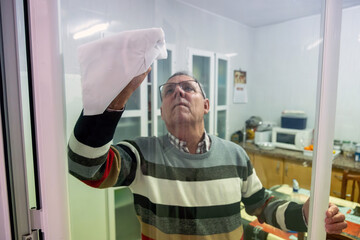 An elderly man wipes a large glass window with a cloth inside a bright kitchen, illustrating routine household maintenance and cleanliness.
