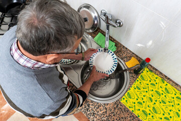 A senior man washes a ceramic plate under running water at the kitchen sink, performing a routine household cleaning task from an overhead view.