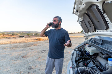 A man stands next to a camper van with the hood open, talking on his phone and gesturing as he seeks assistance in a remote rural area at sunset.