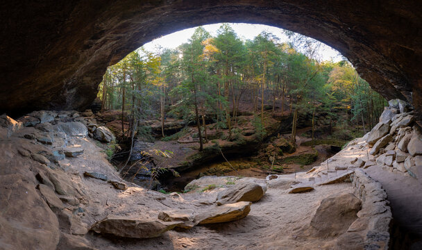 View of a cavernous opening framing a vibrant forest, sunlight dancing on stone steps and rugged rock formations, a natural amphitheater of earthy tones, Logan, Ohio, United States.