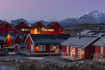 View of red wooden houses glow warmly against the twilight sky, with snow-capped mountains rising in the background, Leknes, Nordland, Norway.
