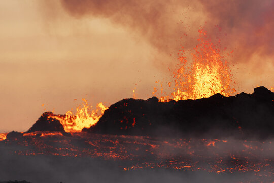 View of molten lava flows like fiery rivers across the dark landscape, erupting with a burst of orange flame against a smoky sky, Reykjanes, Grindavíkurbaer, Iceland.