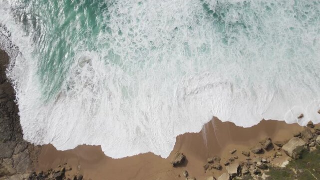 Aerial view of the dramatic coastline where frothy waves meet the sandy shore, a captivating contrast of textures, Costa da Caparica, Setubal, Portugal.