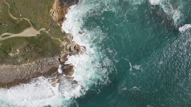 Aerial view of the rugged coastline where the green cliff meets the turquoise sea with white waves crashing, Costa da Caparica, Setubal, Portugal.