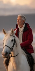 Obraz premium Elderly caucasian male riding horse on beach at sunset in red jacket