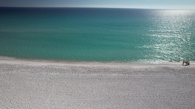 White sand dunes meet the emerald Gulf of Mexico under a bright blue sky. A wide shot captures beachgoers enjoying the clear water and sunny, pristine shoreline.