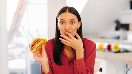 Portrait of happy lady eating tasty juicy burger, licking finger and looking at camera, woman...