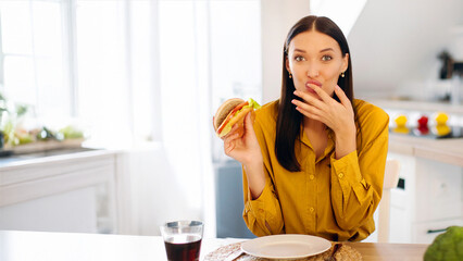 Portrait of happy lady eating tasty juicy burger, licking finger and looking at camera, woman...