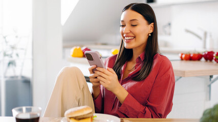 Happy lady sitting at kitchen table, using cellphone while having cheat meal, burger on plate, cola...