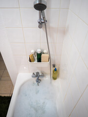 Bathtub filled with foam in a bright tiled bathroom. A faucet, shower head and shelf with bath products create a scene of relaxation and home self-care.