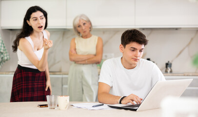 Fototapeta premium Young guy while working on laptop listens to claims of elderly mother and young sister girl in incompetence, unsuccessful study results