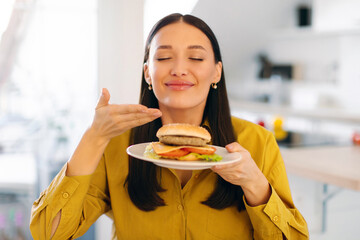 Satisfied woman enjoy fast food snack, smelling burger with closed eyes, being very hungry after hard working day, sitting in kitchen. Eating concept