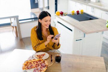 Happy lady using smartphone while eating pizza, writing feedback to delivery service, sitting at...