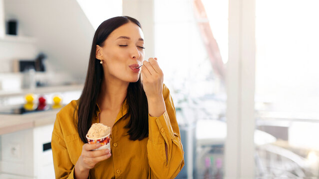 Fototapeta Happy young woman enjoy eating tasty ice cream with closed eyes, sitting in kitchen interior at home, free space