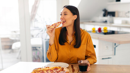 Young satisfied woman eating Italian pizza, enjoying cheat meal at home, sitting at table in kitchen. Yummy unhealthy food
