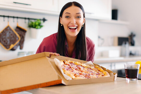 Food delivery concept. Happy European lady holding open box with pizza and looking at camera, standing in kitchen interior