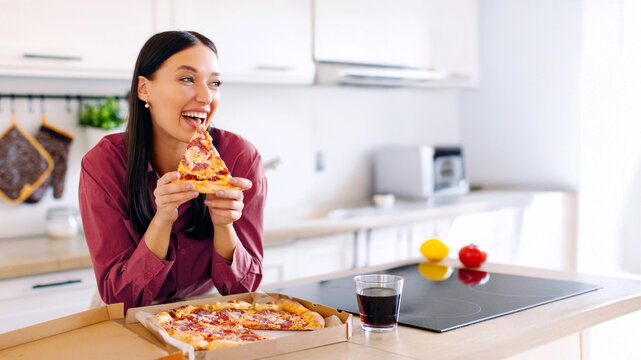 Excited lady holding slice of tasty pizza, enjoying her cheat meal, standing at kitchen counter with carton box from delivery service