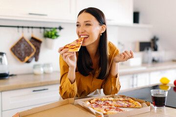 Happy woman biting slice of tasty pizza, enjoying cheat meal dinner at home, sitting at table in...
