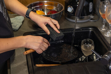 Person washing kitchen spatula under running water. Close-up of hands cleaning a kitchen spatula with a sponge under running tap water in a domestic kitchen sink.