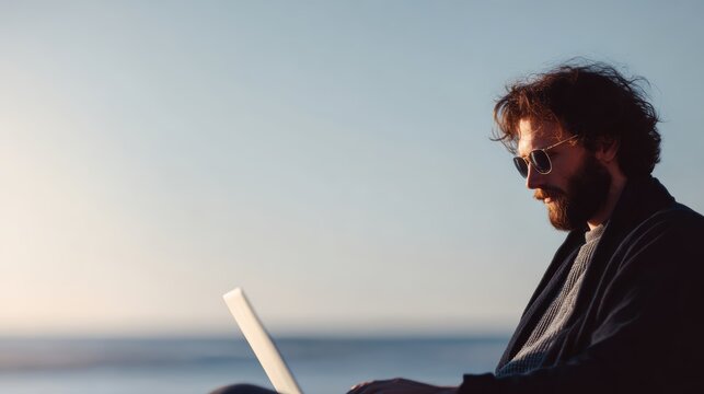 Pensive man freelancer in sunglasses typing on laptop by the sea, remote work vacation concept