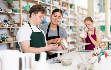 Young female teacher teaching teenage boy and girl students how to paint ceramic cup in ceramic workshop