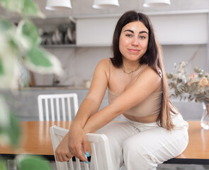 Positive young woman posing cheerfully sitting on table in the kitchen with white furniture