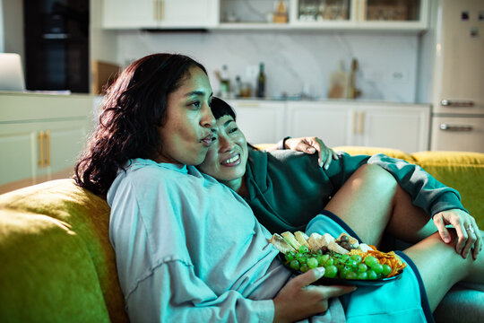 Adult lesbian couple cuddling with snacks at home, relaxed