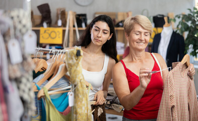Mature woman choosing a warm sweater while standing next to a young Armenian woman who is choosing summer clothes. Two women shopping in a clothing store