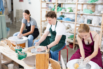 Young woman teacher in apron shows students teenage girl and teenage boy how to sculpt ceramic...