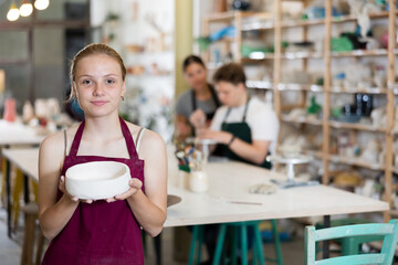 Girl teenager student in apron posing with ceramic bowl in her hands in ceramic workshop