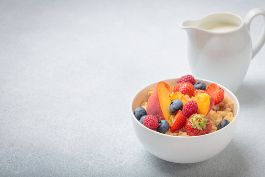 Healthy breakfast with oatmeal porridge with peach, strawberry, raspberry, blueberry and milk in white bowl on gray background, perfect for a nutritious and energizing start to the day, copy space