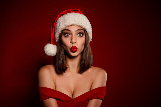 Playful holiday portrait of a woman in Santa hat and red dress making a kissy face against a deep red background