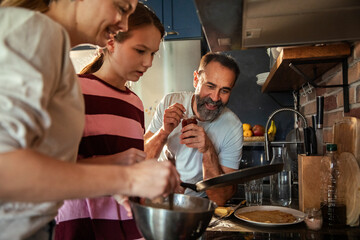 Happy adult parents and teen daughter making pancakes in home kitchen