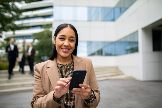 Adult businesswoman smiling with smartphone outside office
