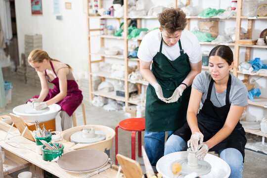 Young woman teacher in apron helps teenager boy and teenager girl students to make product from clay on potter's wheel