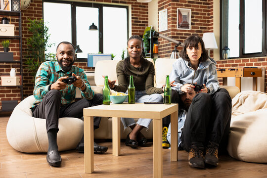 African american man and caucasian woman hold wireless controllers, competing in video game while friends sit nearby watching. Diverse friends enjoy online gaming during weekend hangout in living room