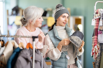 In store, mature woman with young daughter in low customer chooses color and style of winter hat, girl try on headdress. Creating capsule wardrobe, branded clothing at affordable prices.