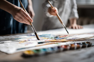 Obraz premium Hands of an adult and child holding paintbrushes, creating colorful artwork on canvas, surrounded by vibrant paints and artistic tools, showcasing a joyful creative process