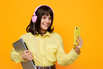 A cheerful young woman wearing pink headphones and a yellow shirt, holding a laptop and smartphone...