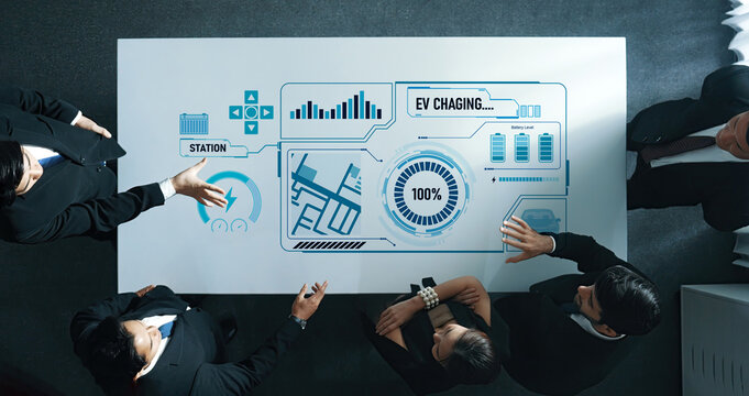 A professional business team collaborates over a modern table, analyzing electric vehicle charging strategies with digital data displays in a contemporary office environment. Trope