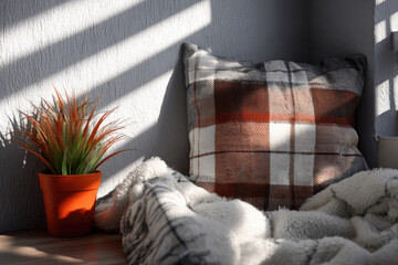 Cozy corner with soft plaid cushion, fluffy blanket, and small indoor plant in sunlight, creating warm and inviting atmosphere for relaxation and comfort