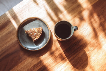 Coffee cup dessert plate wooden table sunlight morning warm shadow breakfast pastry cozy. cup of black coffee and slice of pastry ceramic plate sit sunlit wooden table creating cozy morning