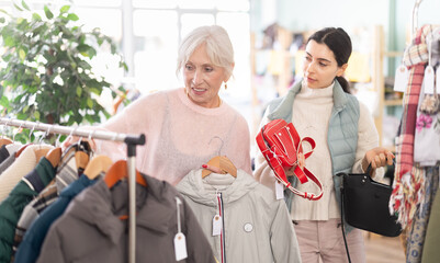 Happy elegance mature woman selecting new coat or jacket for cold season and armenian girl choosing handbag in modern clothing boutique