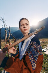 Naklejka premium Woman outdoors in mountains holding a wooden staff across her shoulders, rural portrait at sunrise with patterned jacket and meadow valley background, thoughtful gaze in warm sunlight.