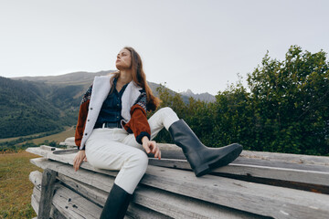 Naklejka premium Woman in nature sits on wooden fence overlooking mountains, outdoor fashion with boots and warm jacket, relaxed pose in meadow landscape for travel and lifestyle.