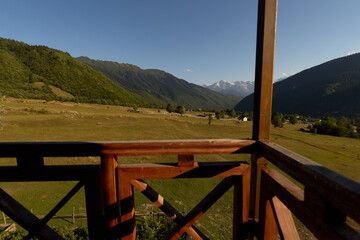 Obraz premium Balcony wooden railing overlooking mountains and valley with green meadow and countryside field, rustic cabin view under clear sky and distant snowy peaks for nature retreat.