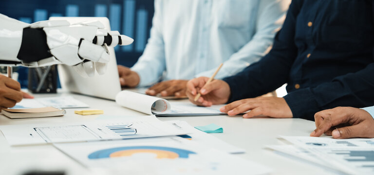 A diverse business team engaged in collaborative work with the assistance of a robotic arm, analyzing data and strategizing in a modern office setup.Noogenesis