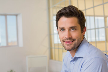 Man sitting at office in blue button-down shirt by large window with glass partition and plant